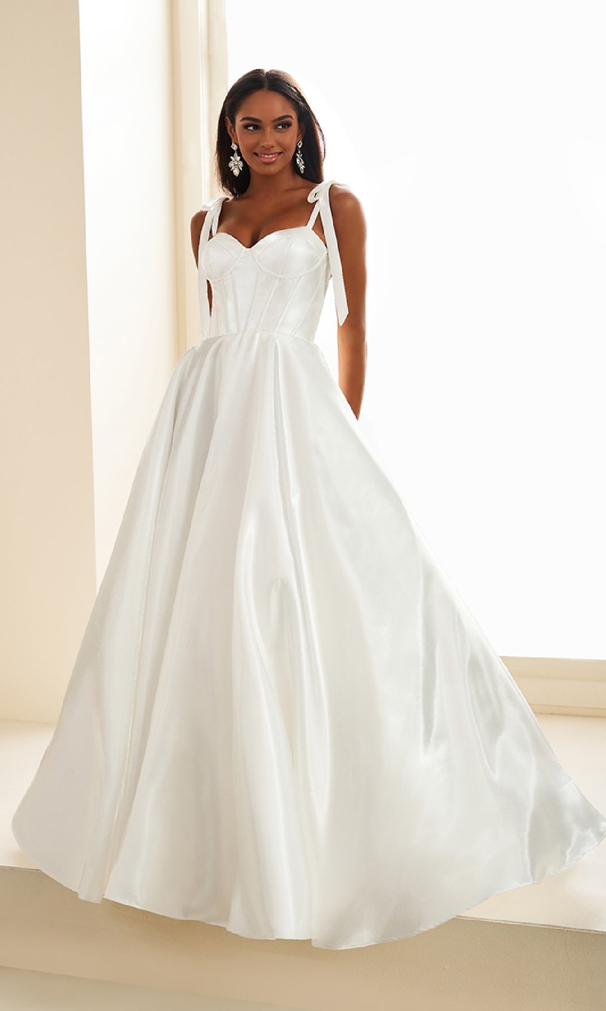 A woman stands indoors near a window wearing the Ellie Wilde EW36210 Shoulder-Bows Prom Ball Gown, a white satin lace-up dress featuring a fitted bodice, bow straps, and a full skirt.